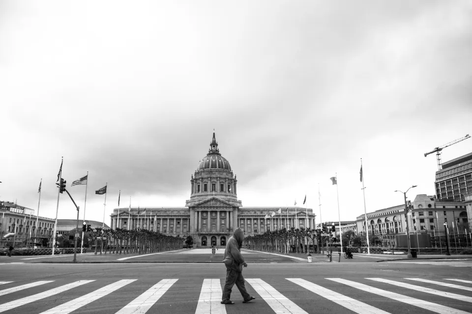 Black and white photo of San Francisco city hall neoclassical dome with person walking across crosswalk in foreground.